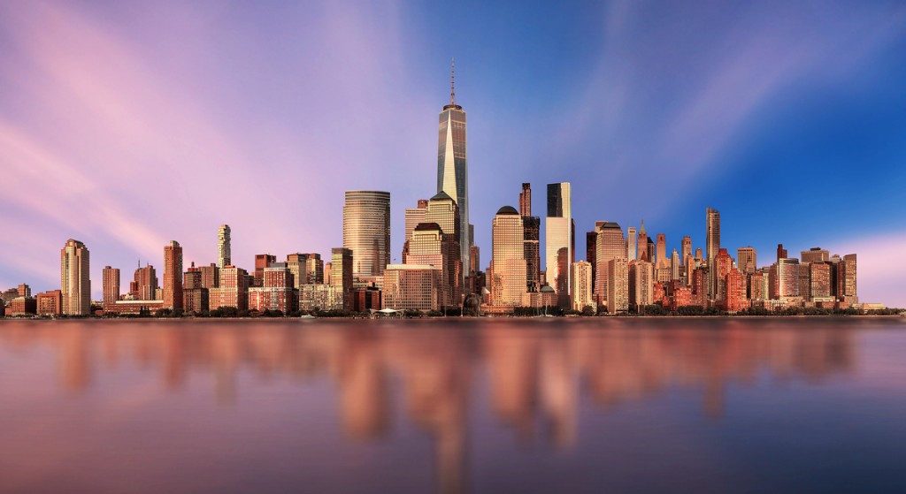 Lower Manhattan skyline across the water at sunset, with One World Trade Center at the center.