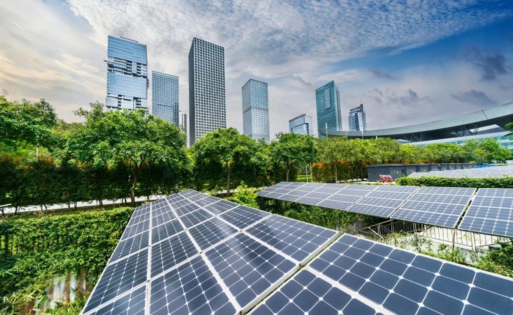 Solar panels in the foreground with a modern glass skyscraper skyline and green trees behind them.