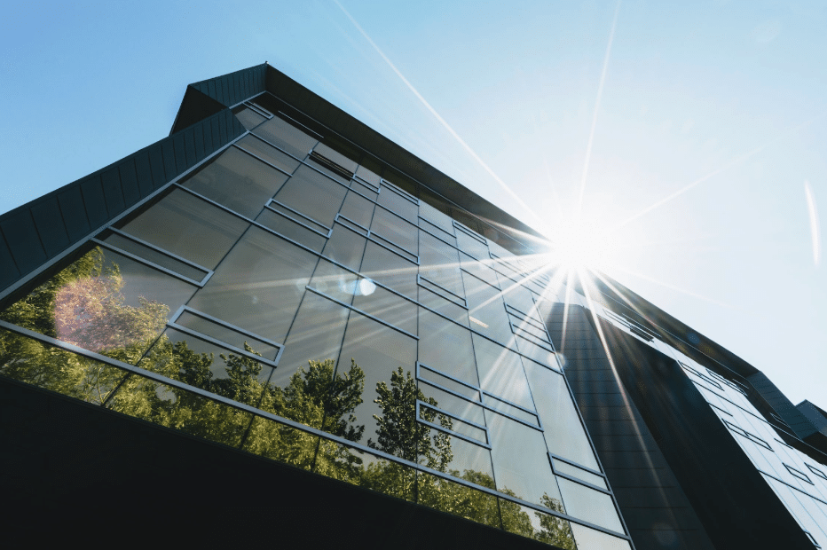 Looking up at a modern glass commercial building under a bright blue sky, with trees reflected in the facade.
